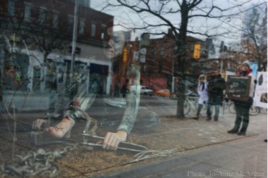 Shannon Kornelsen of Fur-Bearer Defenders in a leg-hold trap with scenes from Queen Street (including a couple wearing fur-trimmed jackets) reflected in the shop window. Photo: JMcArthur/WeAnimals